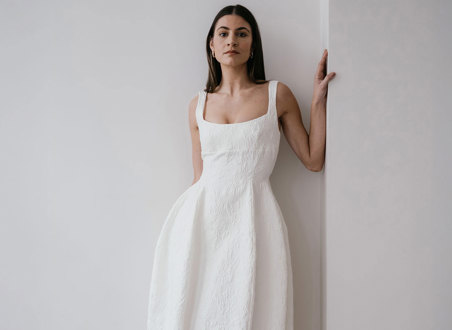 a dark haired model stands in the corner of two plain white walls, leaning to one side, her hand resting on the wall at shoulder height as she wears textured white wedding dress with a structured hourglass shape