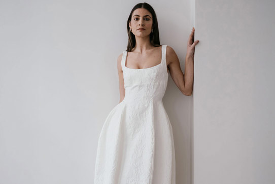 a dark haired model stands in the corner of two plain white walls, leaning to one side, her hand resting on the wall at shoulder height as she wears textured white wedding dress with a structured hourglass shape