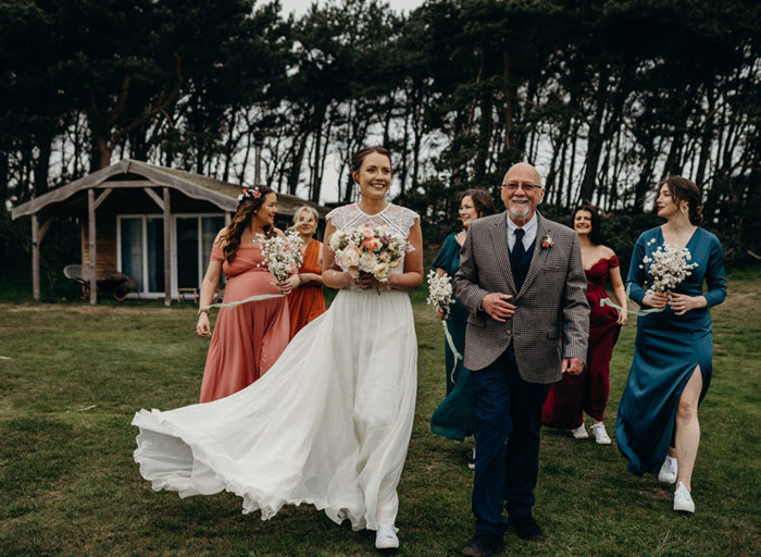 a bride walking across a lawn with a group of people. One is a man wearing a checked suit and the others are wearing bridesmaid dresses in shades of teal, orange, pink and red. The bride is carrying a pastel posy of flowers and there is a small wooden hut with large windows in the background