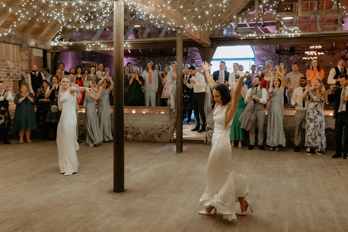 two brides dancing on a wooden floor under festoon string lights as guests cheer on at the Byre at Inchrya wedding reception
