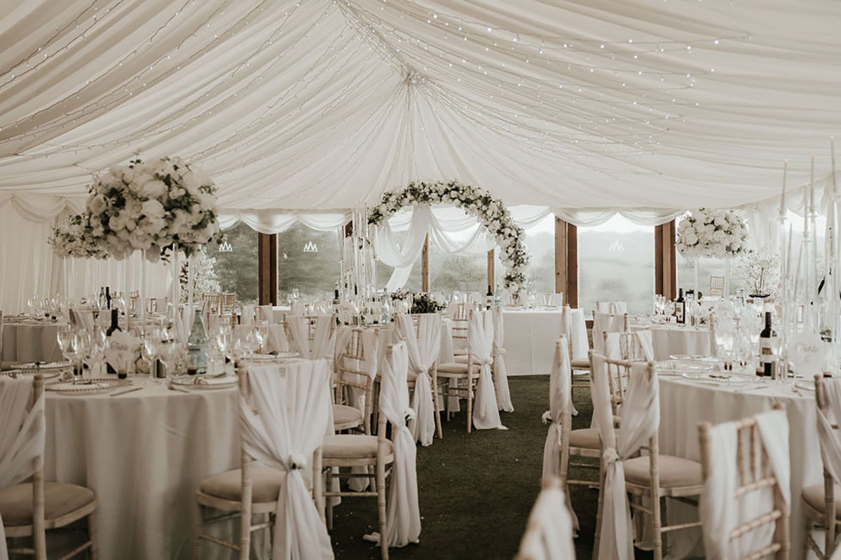 White marquee wedding reception with round tables, tall floral arrangements and draped ceiling lights.