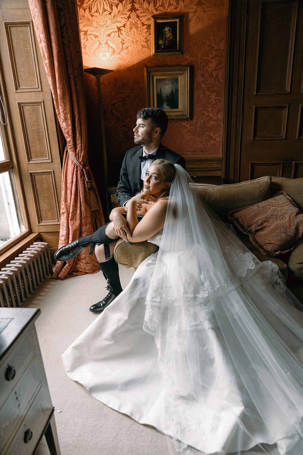 Bride in flowing veil and groom in black-tie kilt look out window of richly decorated room at Dundas Castle.