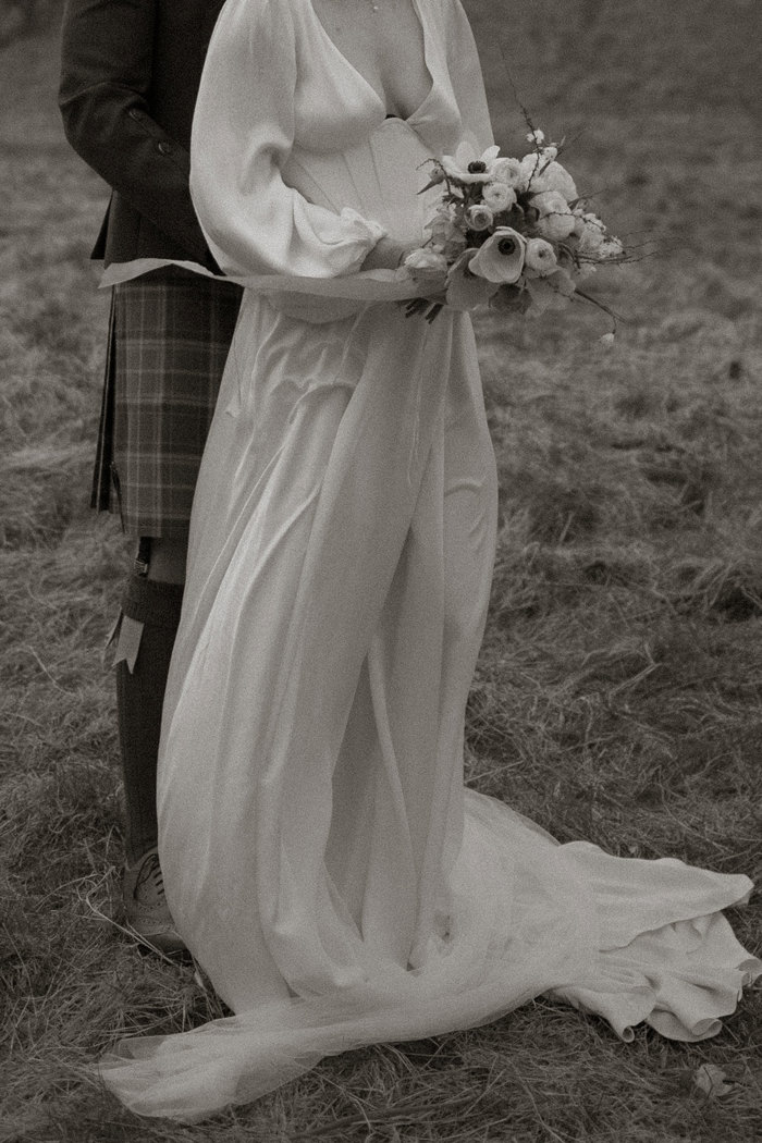 black and white photo of bride in plunging satin dress holding a bouquet of flowers, standing in front of groom in kilt outfit, both captured from the neck down, stood outdoors on the grass