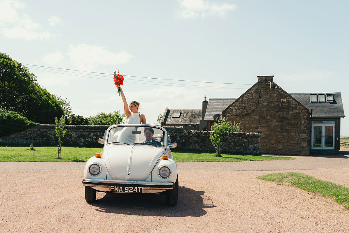 Bride stands in a white vintage Volkswagen Beetle convertible holding her bouquet while arriving at Kinkell Byre wedding venue in St Andrews.
