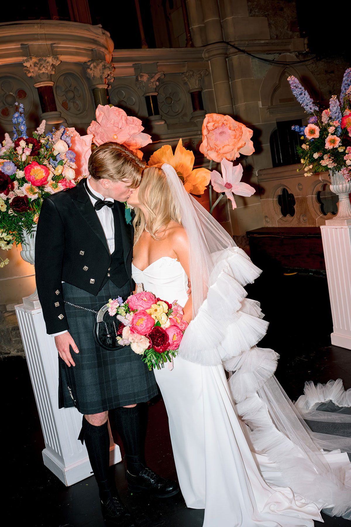 Bride and groom kissing beside oversized paper flowers and colourful floral arrangements during an indoor wedding ceremony setup