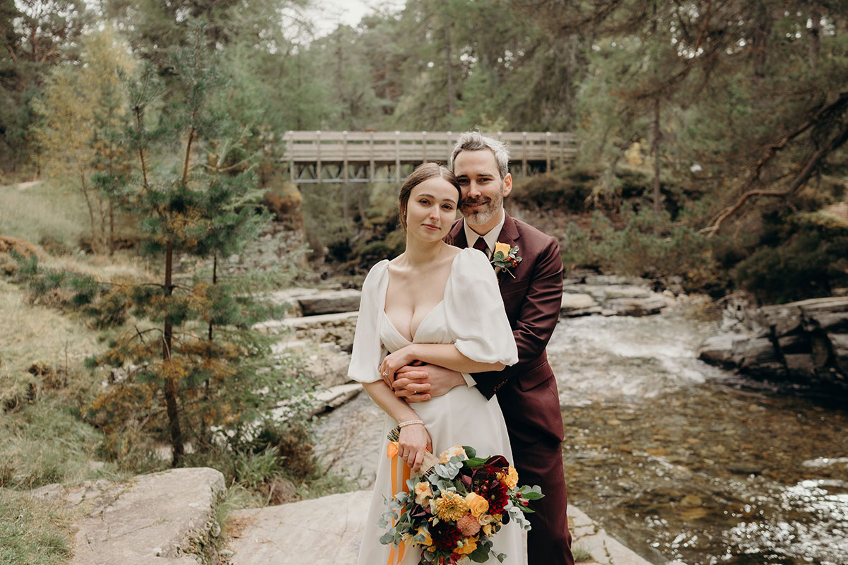 a bride and groom posing by a wooden bridge over the River Quoich on Mar Lodge Estate