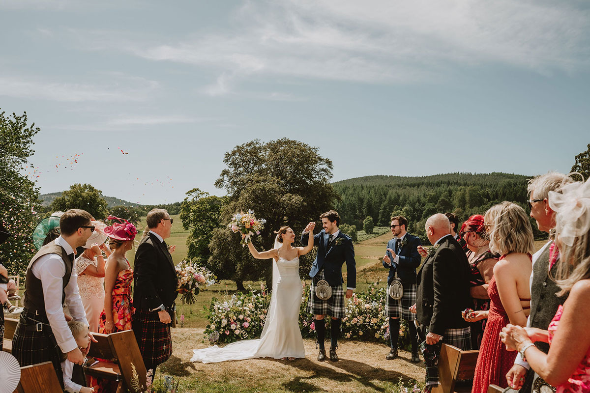 Newly married couple celebrating outdoors as guests throw confetti at a countryside ceremony, with scenic hills and trees in the background
