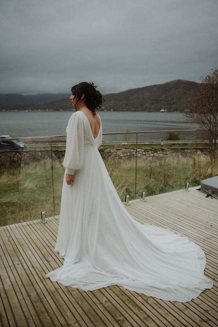 a bride standing on wooden decking on a glass balcony looking out onto a loch and mountains in the Scottish Highlands