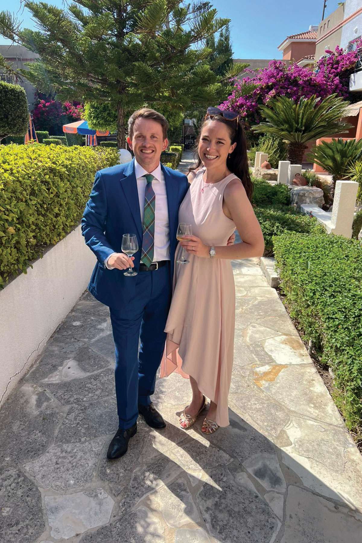 Couple smiling and holding drinks in a sunny garden courtyard with palm trees and bright pink flowers; the man wears a blue suit and tartan tie and the woman wears a pale pink dress