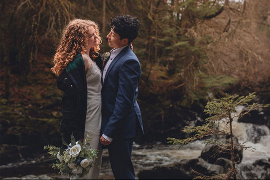A couple looking into each other's eyes while standing next to a river in a forest
