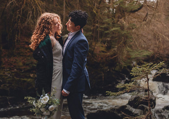 A couple looking into each other's eyes while standing next to a river in a forest
