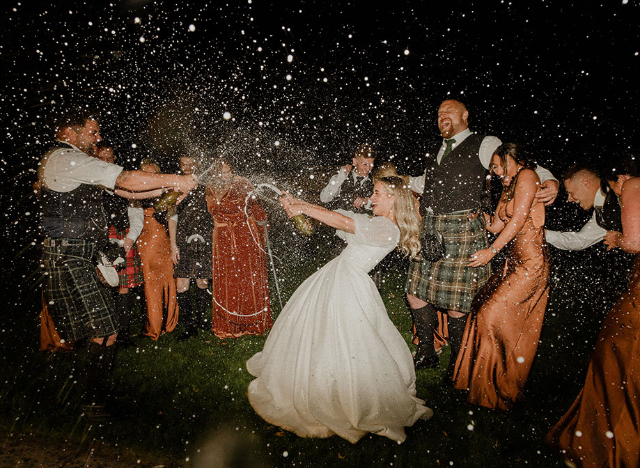 a bride and groom doing a champagne spray at each other in the dark with men in kilts and women in rust coloured bridesmaid dresses cheer and try to avoid spray in the background