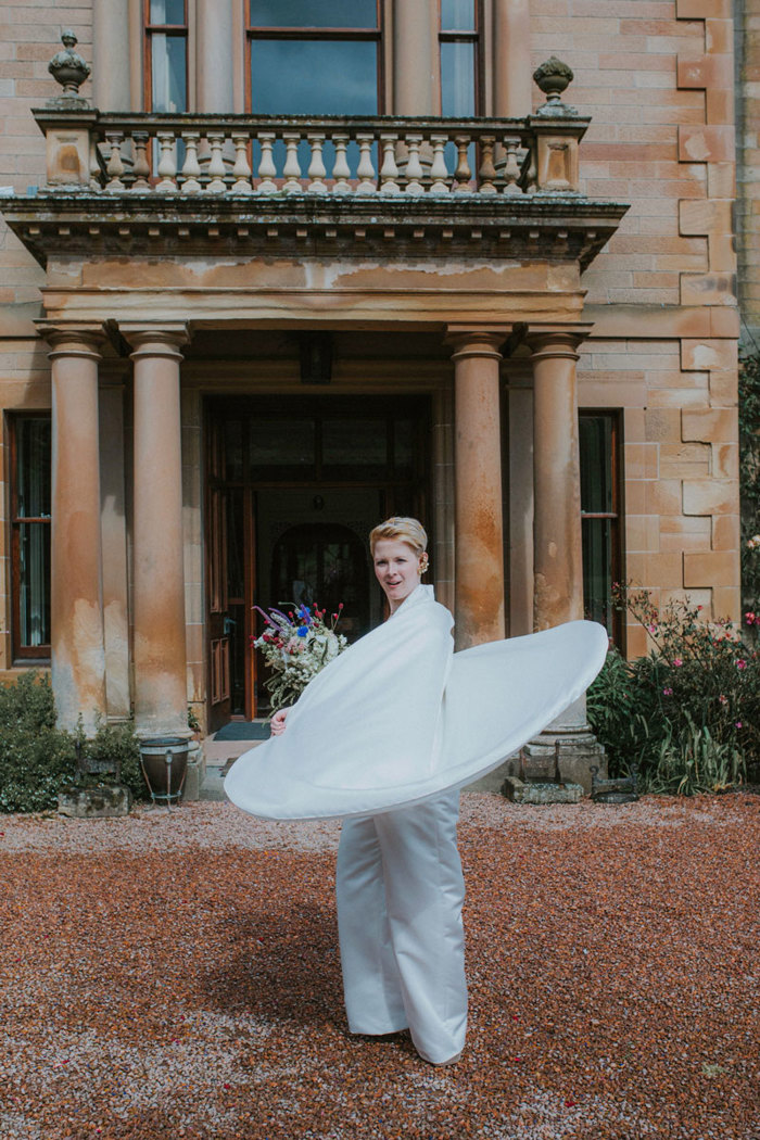 bride holding a wildflower bouquet twirls custom made wedding cape outside cambo house