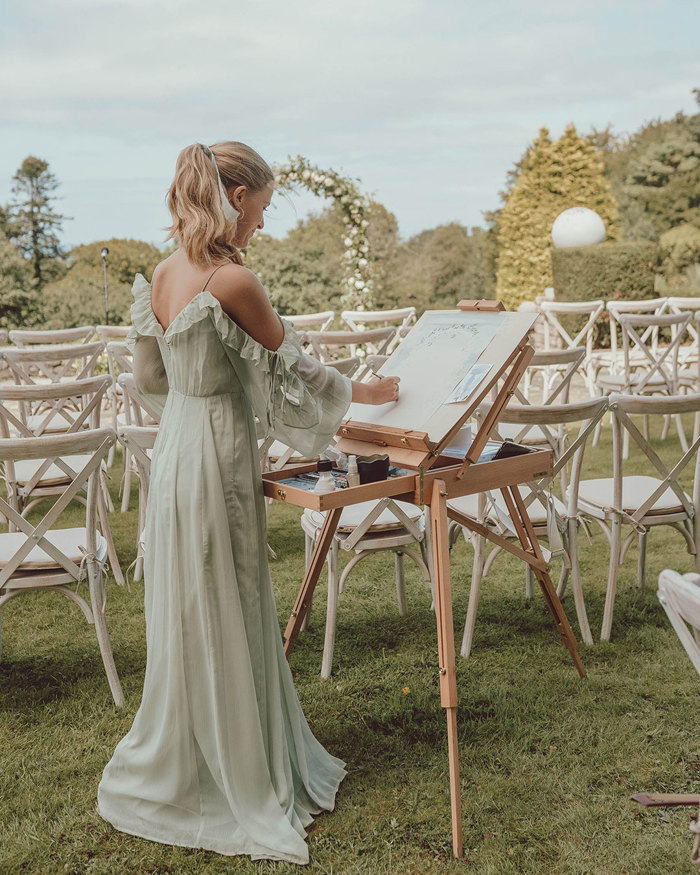 a blonde woman in a floaty green dress stands at an easel, painting a wedding ceremony set up