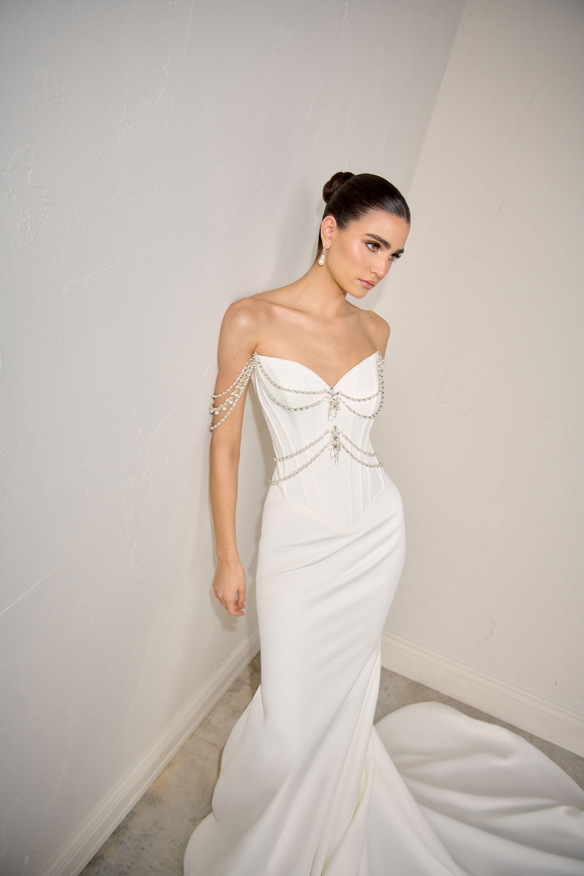 Model in a sleek strapless wedding dress with structured corset bodice and delicate draped chain detailing across the shoulders and waist, photographed against a neutral studio wall