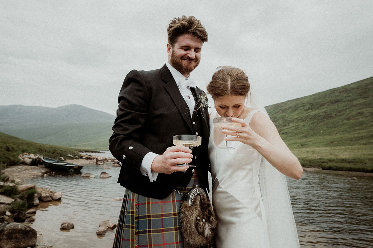 bride and groom drink from champagne coupes while standing next to a river with misty mountains behind them