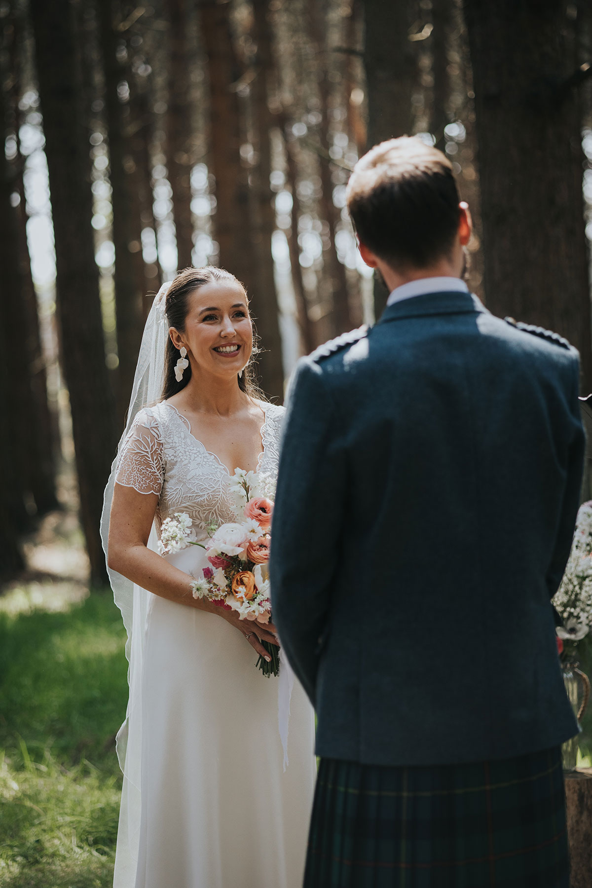 bride smiles during wedding ceremony while wearing a veil and holding colourful flowers as the groom is out of focus