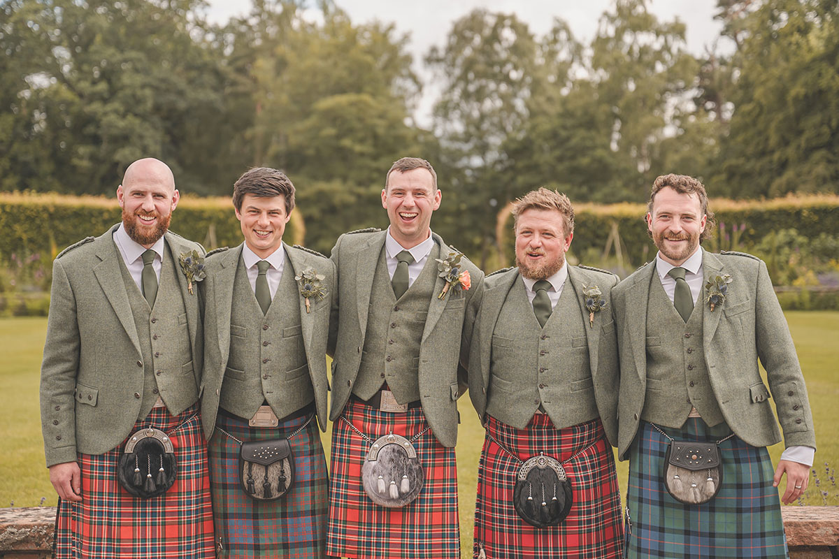 Five men stand with their arms around each other smiling in a green jackets and different coloured kilts