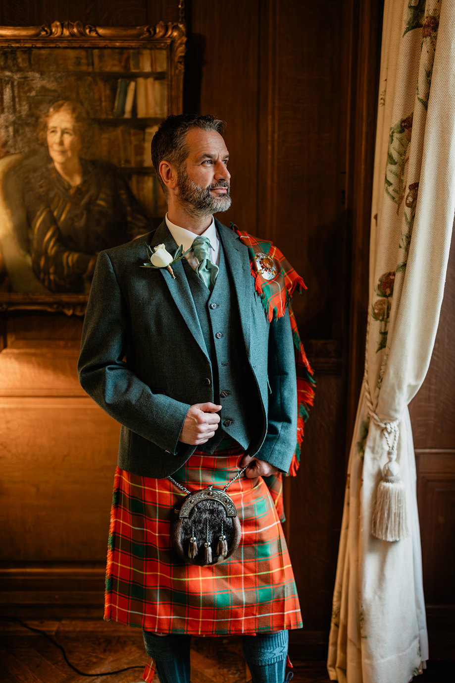 groom dressed in a red and green tartan kilt outfit, holds onto his jacket with one hand and sporran with the other while casting a look out the window