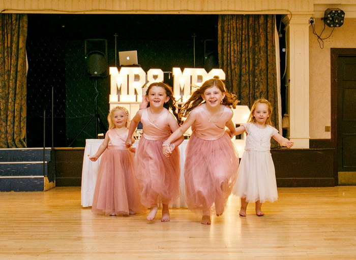 Three flower girls in pink dresses and one girl in a white dress run across a dancefloor towards the camera and away from a light up 'Mr & Mrs' sign