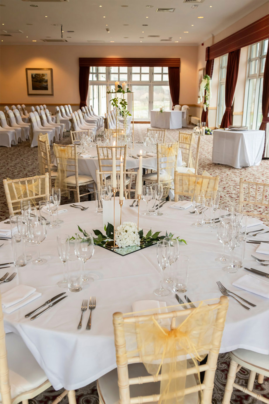 A room set up for a wedding ceremony on one side and a wedding reception on the other side with wooden chairs that have yellow ribbons tied around them, round tables with white tablecloths