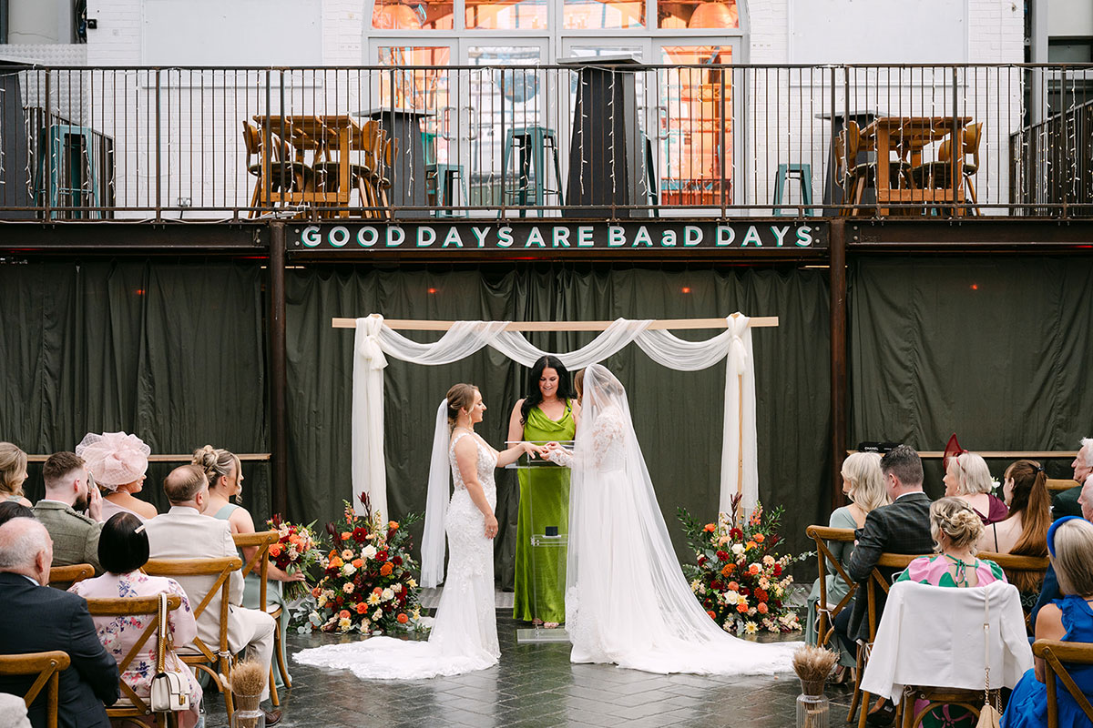 Wedding ceremony at BAaD Glasgow with two brides exchanging rings beneath a simple draped ceremony arch.