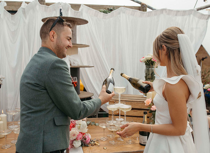 a bride and groom pouring champagne into a small tower of champagne coupes that are sitting on a wooden surface. There is a white curtain in the background