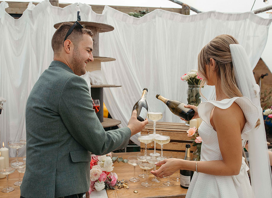 a bride and groom pouring champagne into a small tower of champagne coupes that are sitting on a wooden surface. There is a white curtain in the background