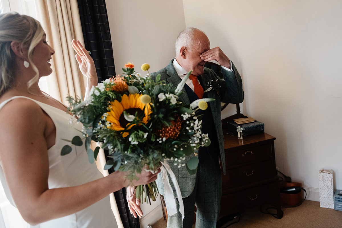 motional father wiping tears as he sees the bride holding a vibrant sunflower bouquet before the ceremony