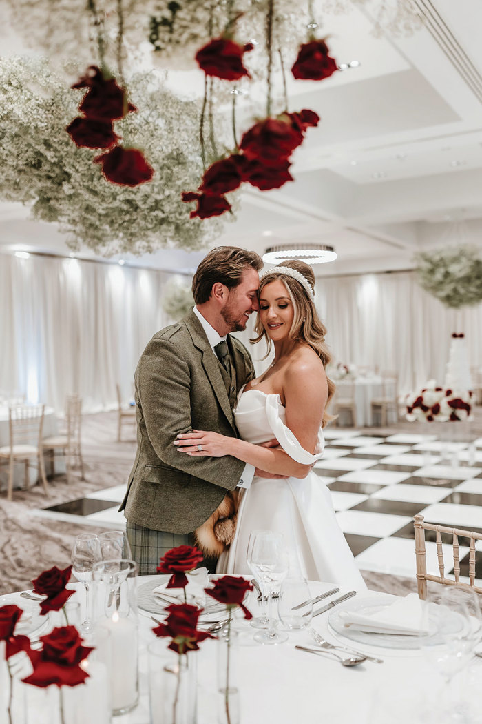 bride and groom stand chest to chest, the groom's nose leaning into her cheek, both smiling and surrounded by various displays of red roses in a black and white dining room