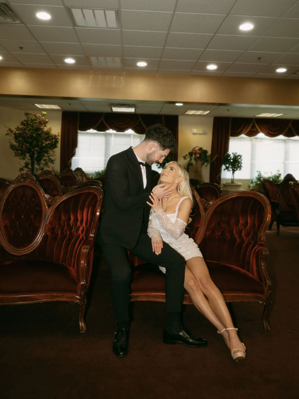 bride and groom look adoringly at each other whilst sat in a line of plush red velvet chairs