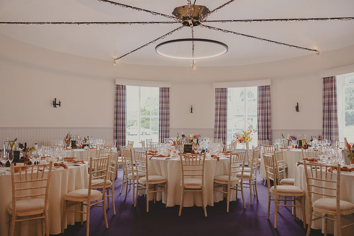 Bright circular dining room with round tables set for a wedding meal, featuring cream linens, floral centrepieces, and lilac tartan curtains