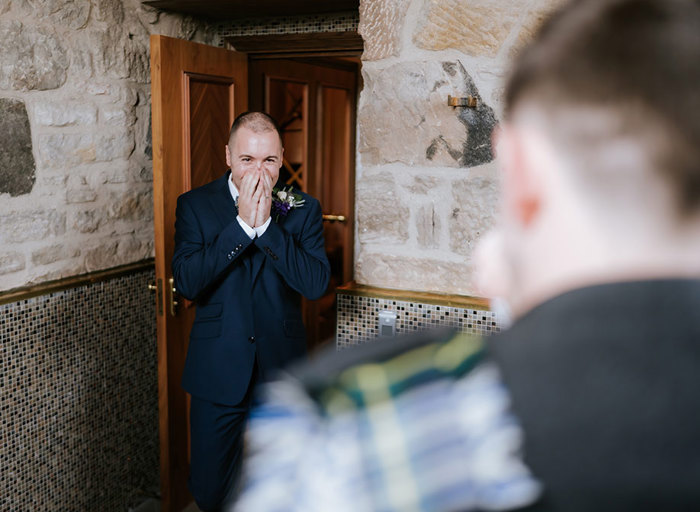 a groom holding his hands up to his face in happy surprise as he looks at other groom during a first look