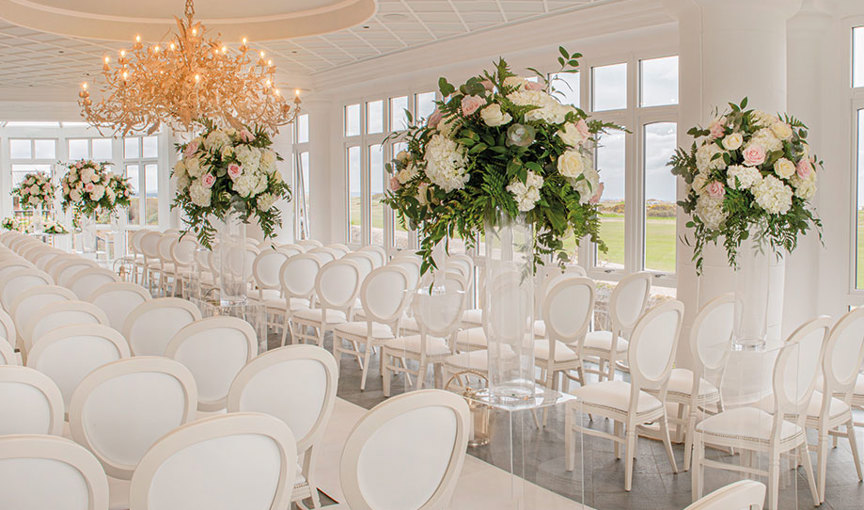 Interior of conservatory at Old Course Hotel set for a wedding ceremony with elegant white chairs, white aisle runner and large flower arrangements on perspex plinths