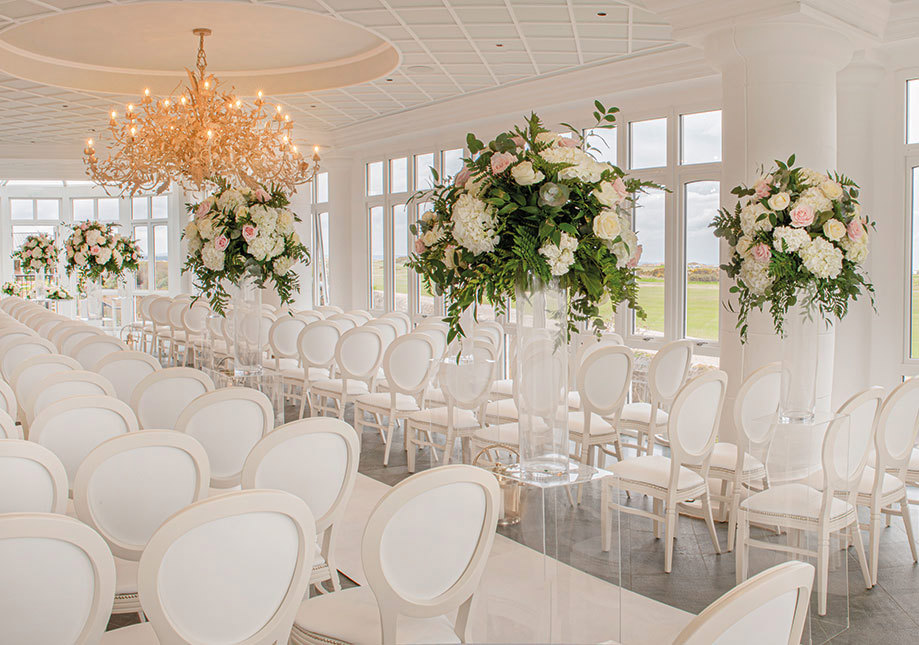 Interior of conservatory at Old Course Hotel set for a wedding ceremony with elegant white chairs, white aisle runner and large flower arrangements on perspex plinths