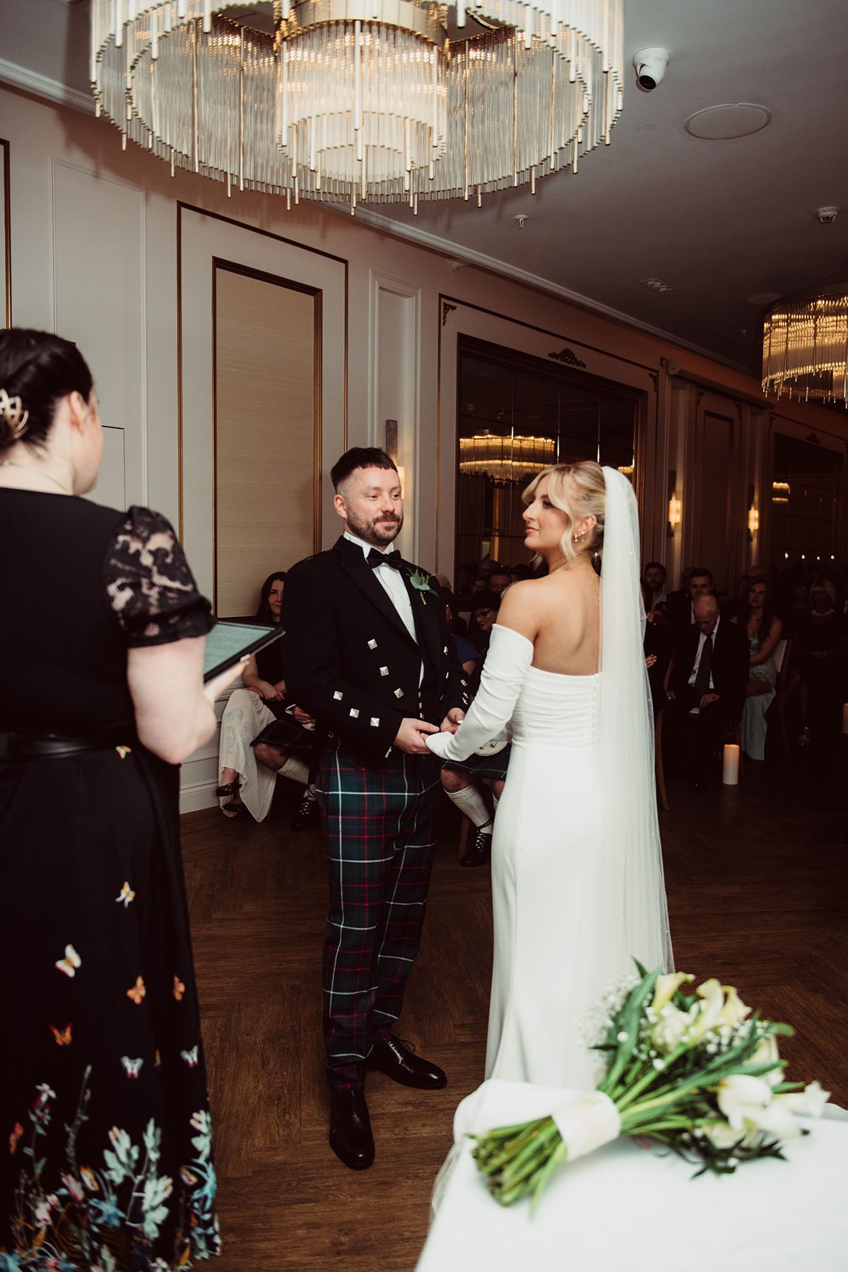Bride and groom holding hands during a candlelit indoor ceremony beneath a large chandelier, with guests seated around them