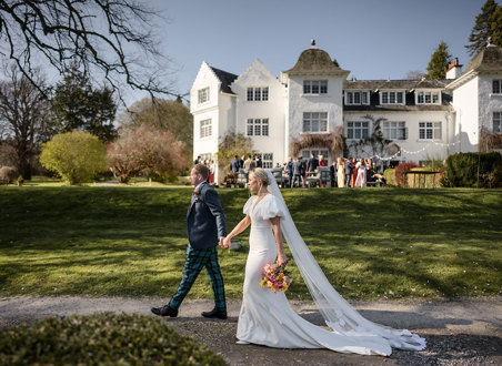 bride wearing a veil walks hand in hand with groom in front of achnagairn castle with guests pictured in the background
