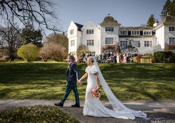 bride wearing a veil walks hand in hand with groom in front of achnagairn castle with guests pictured in the background