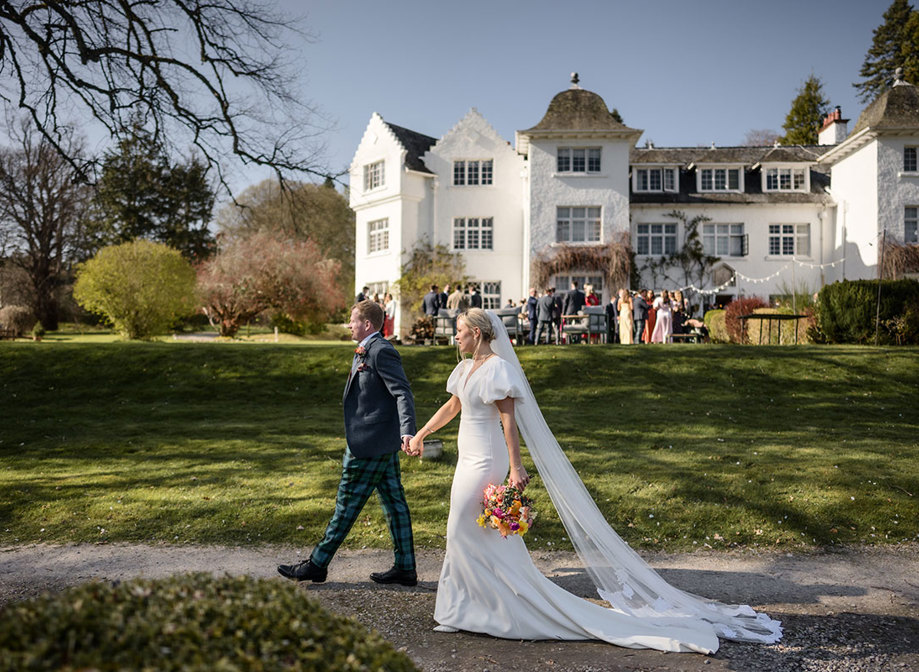 bride wearing a veil walks hand in hand with groom in front of achnagairn castle with guests pictured in the background