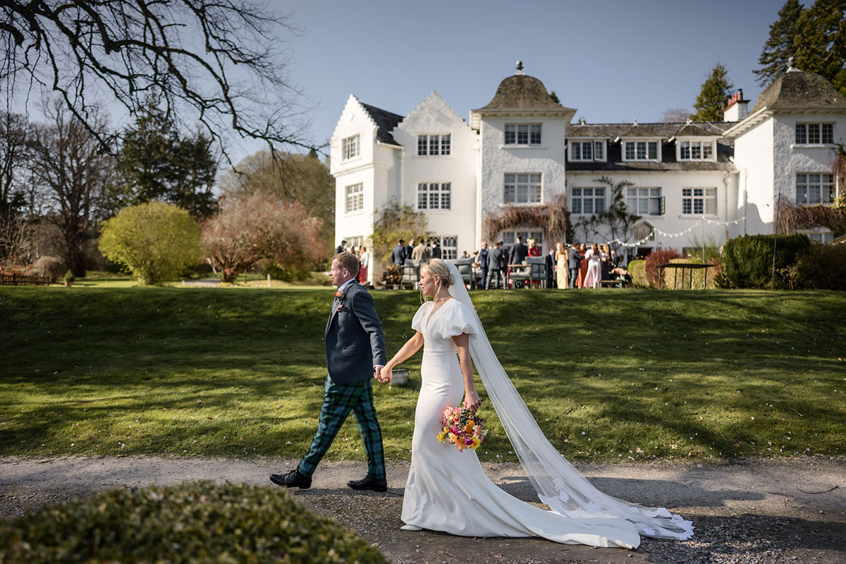 bride wearing a veil walks hand in hand with groom in front of achnagairn castle with guests pictured in the background