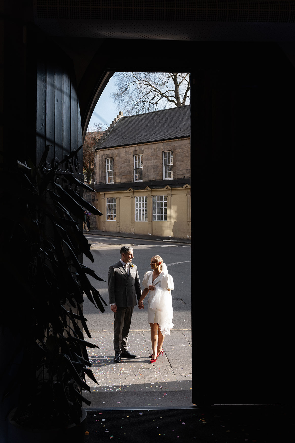 Bride and groom holding hands outside Edinburgh City Chambers, photographed through an arched doorway with confetti scattered on the ground.