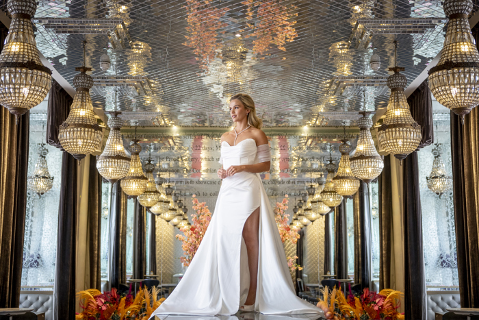 A woman wearing a strapless wedding dress with tulle sleeves and a slit in the skirt standing in a mirrored room with crystal chandeliers