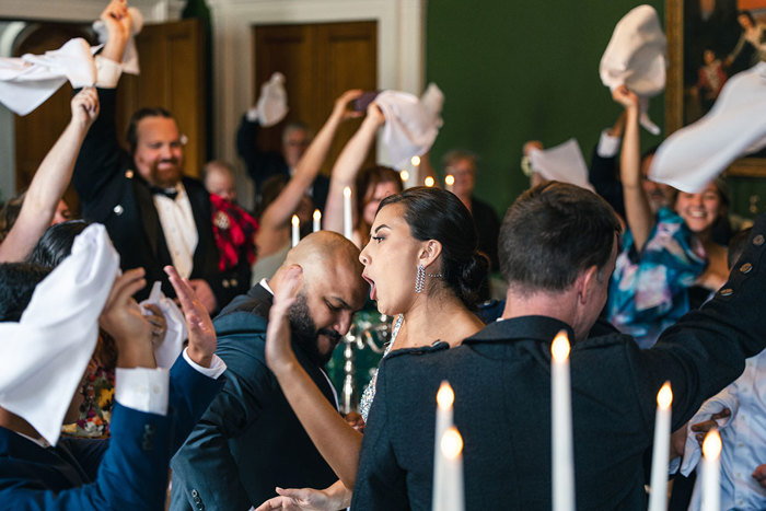 people wave white napkins as a person wearing a silver glitter dress walks through a room at Blairquhan Castle with white tapered candles in foreground