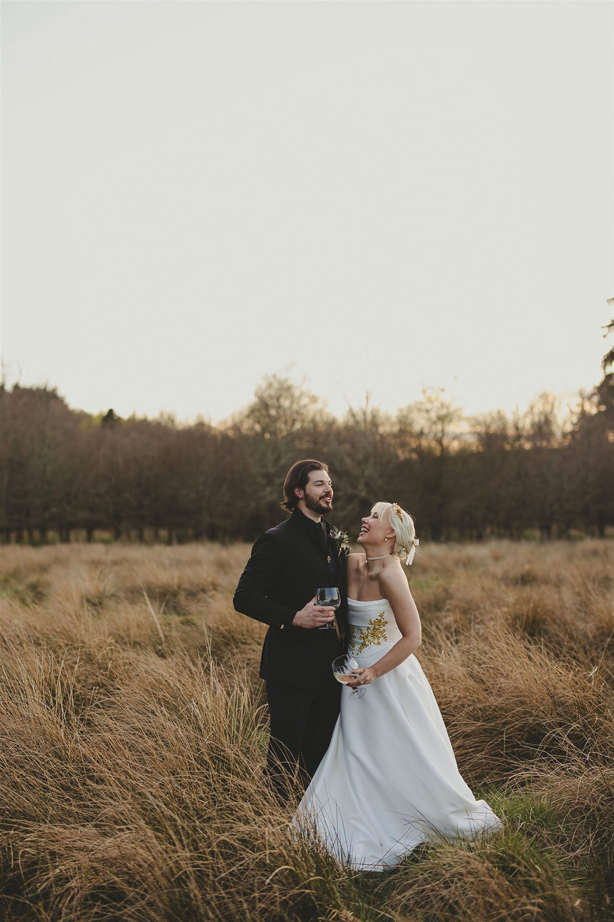 Bride and groom laughing together while standing in a grassy field at sunset, holding glasses and enjoying a relaxed moment outdoors.
