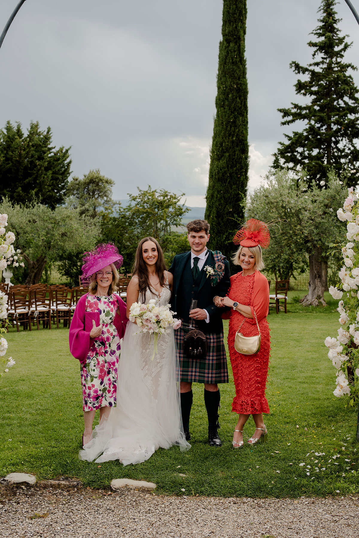 bride in flowy tulle wedding dress stands alongside groom in tartan kilt outfit, her mum by her side wearing pink floral midi dress with matching pink hat, while groom's mum wears orange midi dress with matching orange percher hat