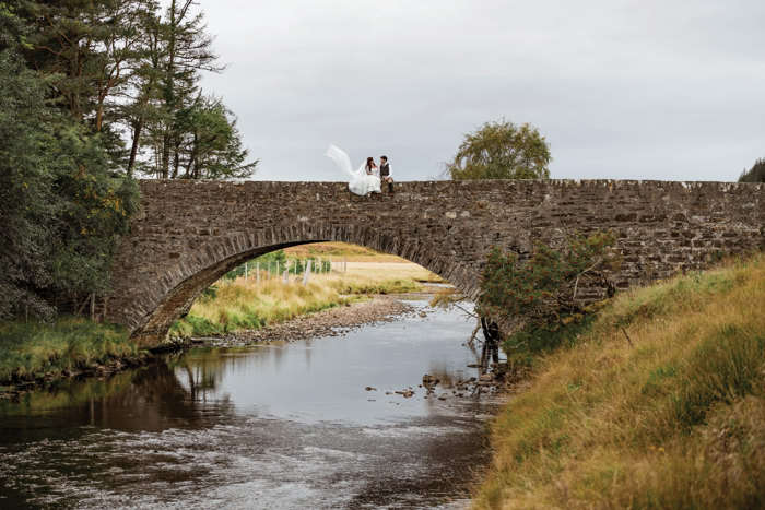 bride and groom sit on bridge with veil flying in the wind