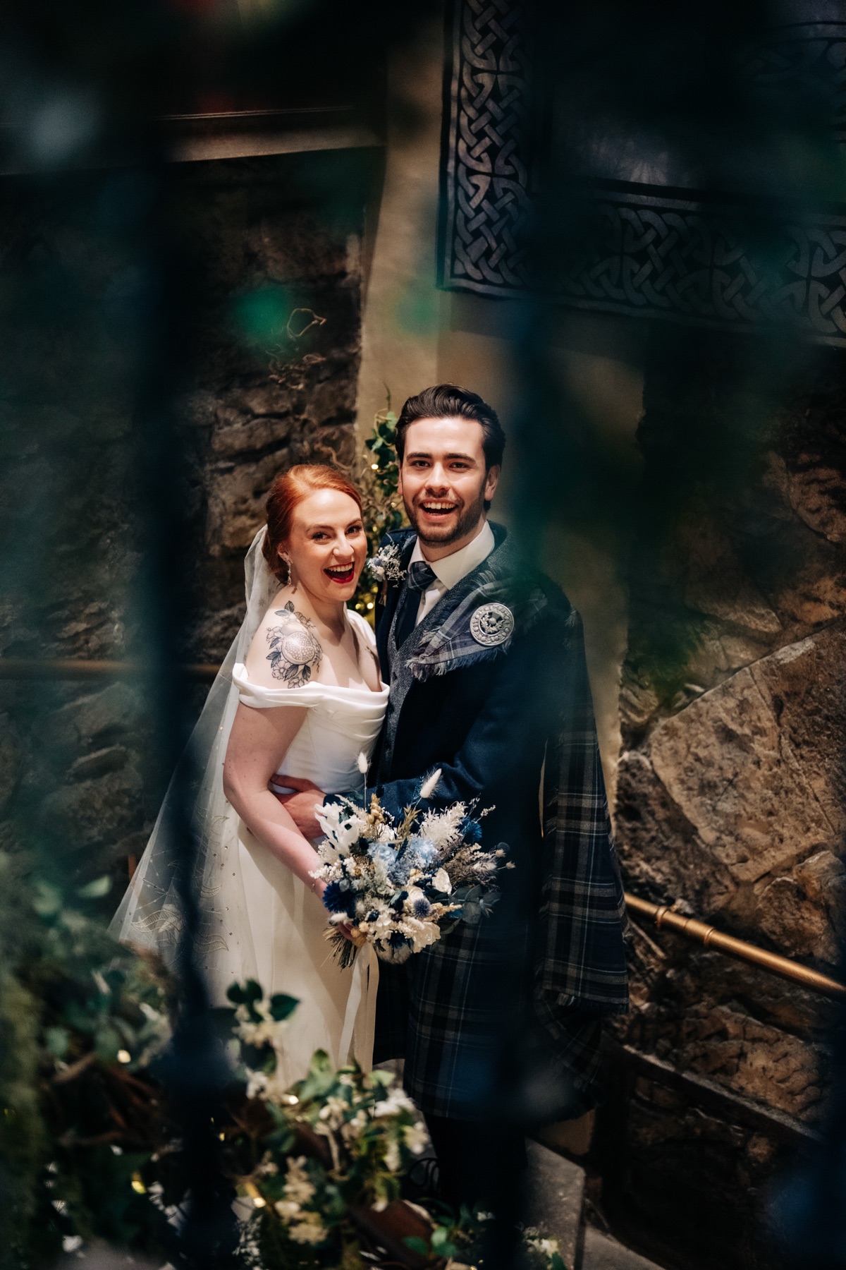 Robyn and James pose for a wedding portrait on the stone staircase at Òran Mór in Glasgow, with Robyn holding her blue-and-white bridal bouquet.