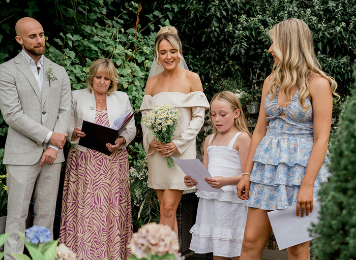 a young girl reading from a sheet of paper in a garden as a line up of bride, groom, celebrant and person wearing a blue flower dress look on