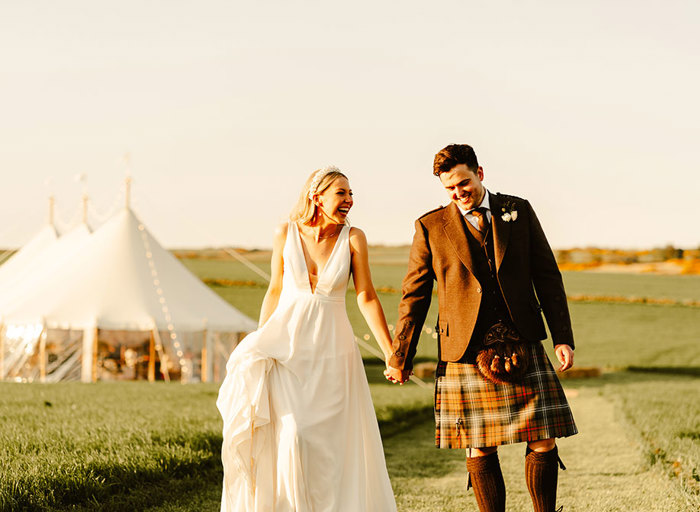 a bride and groom holding hands walking through a golden sunlit field with marquee in the background
