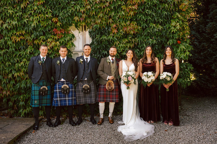 A groom and groomsmen in different colours kilts, a bride in a white dress and bridesmaids in burgundy velvet dresses stand in front of a leafy wall